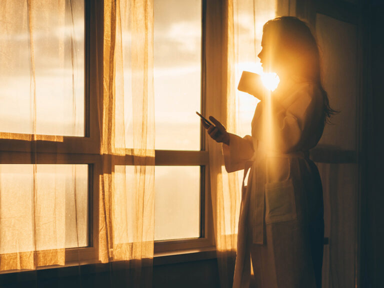 A client watches the beach sunrise from a window while drinking coffee and checking their Amerant account on a phone.