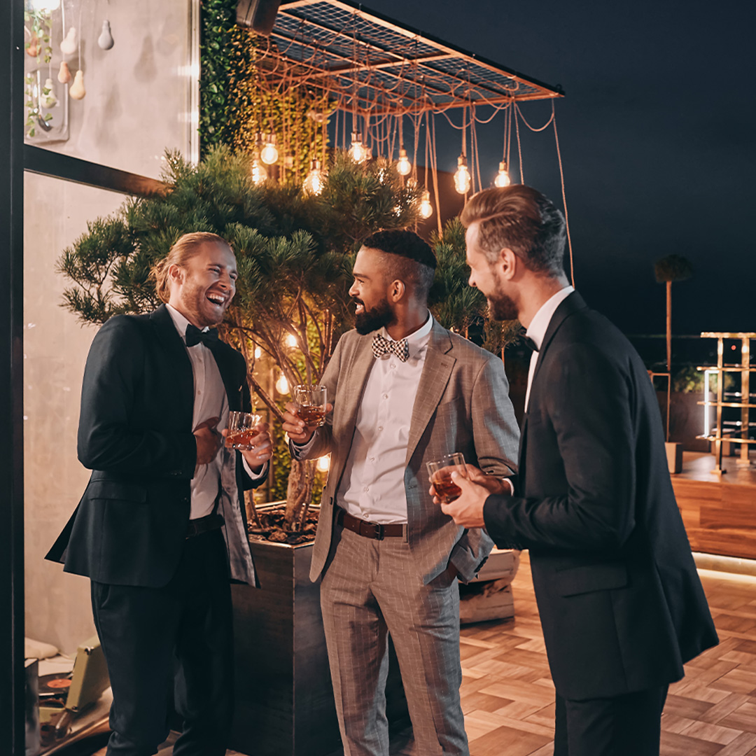 Three men laughing wearing formal attire on a rooftop with drinks.