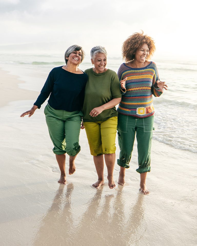 three older-aged friends walk on the beach smiling